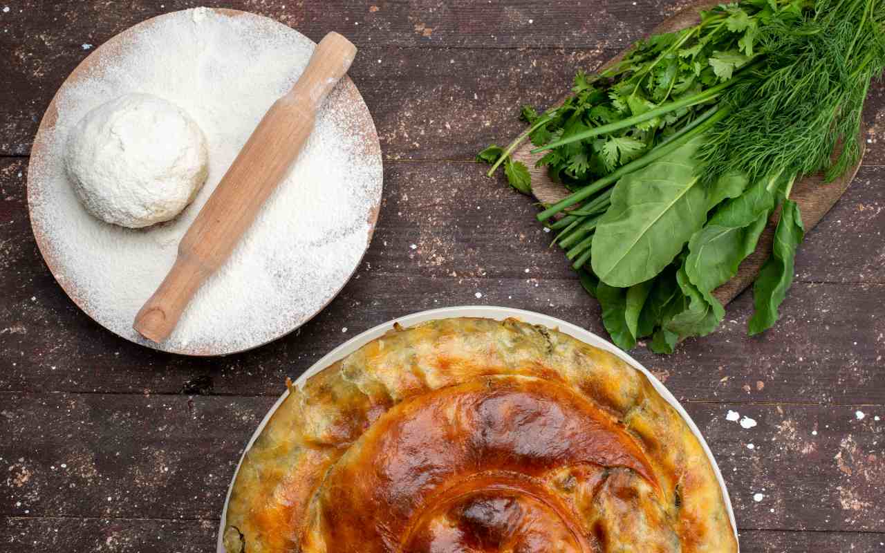 Una torta di scarola accanto a un impasto salato e verdure fresche su un tavolo