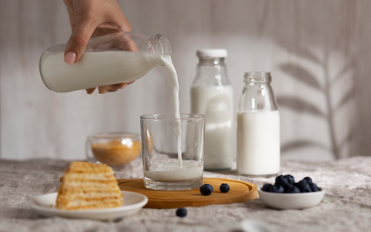 Una persona versa del latte in un bicchiere accanto a bottiglie di latte, dolci e frutta